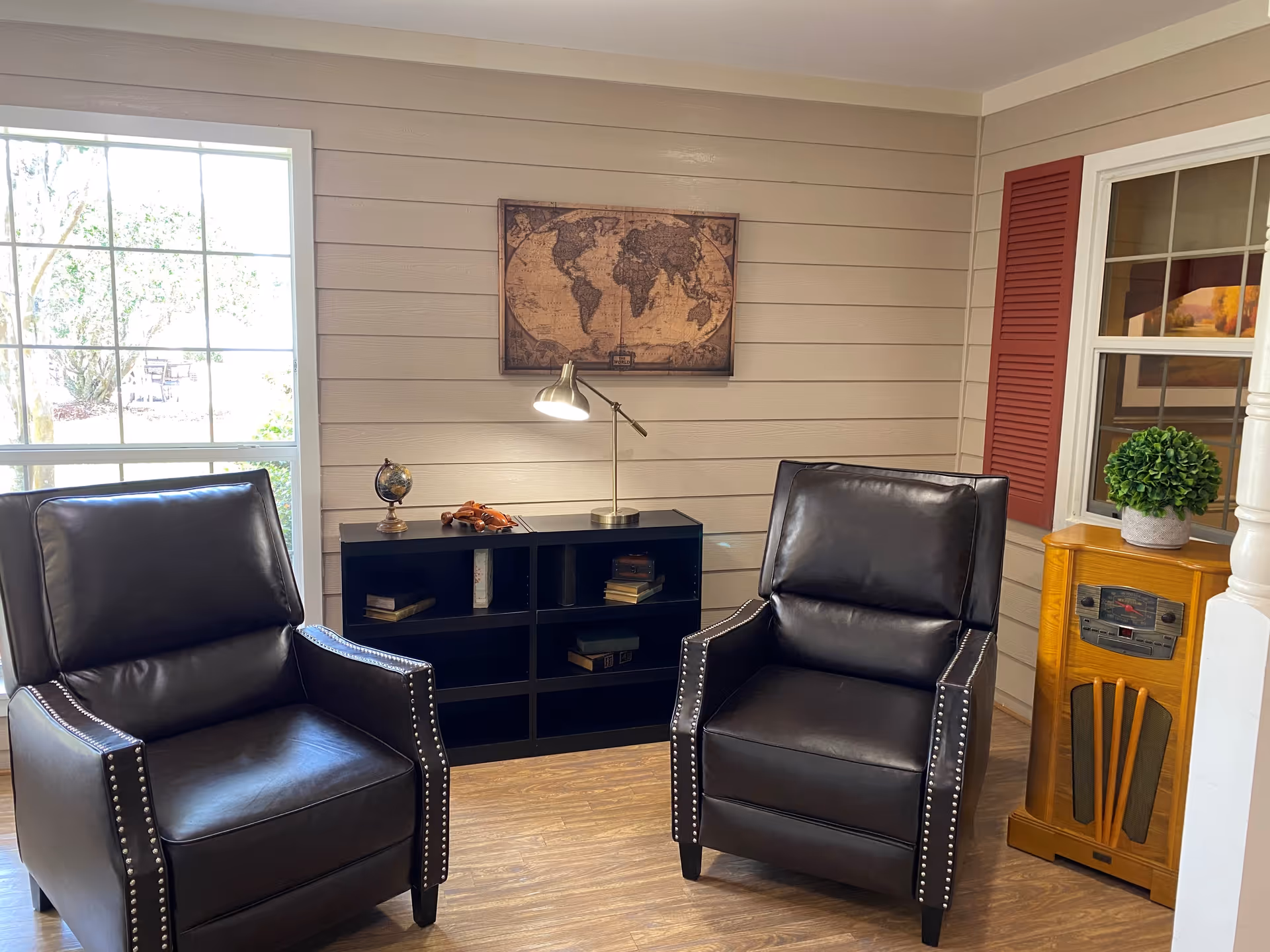Two dark leather armchairs in a cozy living area with a wall map, black shelving, lamp, window, and decorative accents.