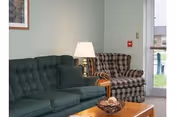 A cozy living room area with a green tufted sofa and a plaid armchair. A wooden coffee table with a decorative bowl is in front of the seating. A table lamp is placed between the sofa and armchair. A framed picture hangs on the light green wall, and a glass door is visible in the background.