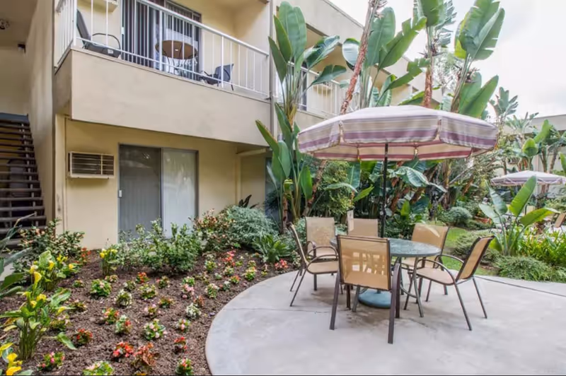 Outdoor patio area with a round glass table and six chairs under a striped umbrella, surrounded by lush green plants and flowers. The patio is adjacent to a two-story building with balconies and sliding glass doors.