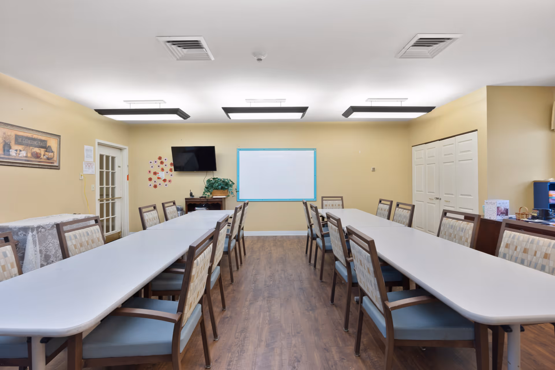Long multipurpose dining/activity room with two rows of tables and chairs facing a whiteboard and a wall-mounted TV.