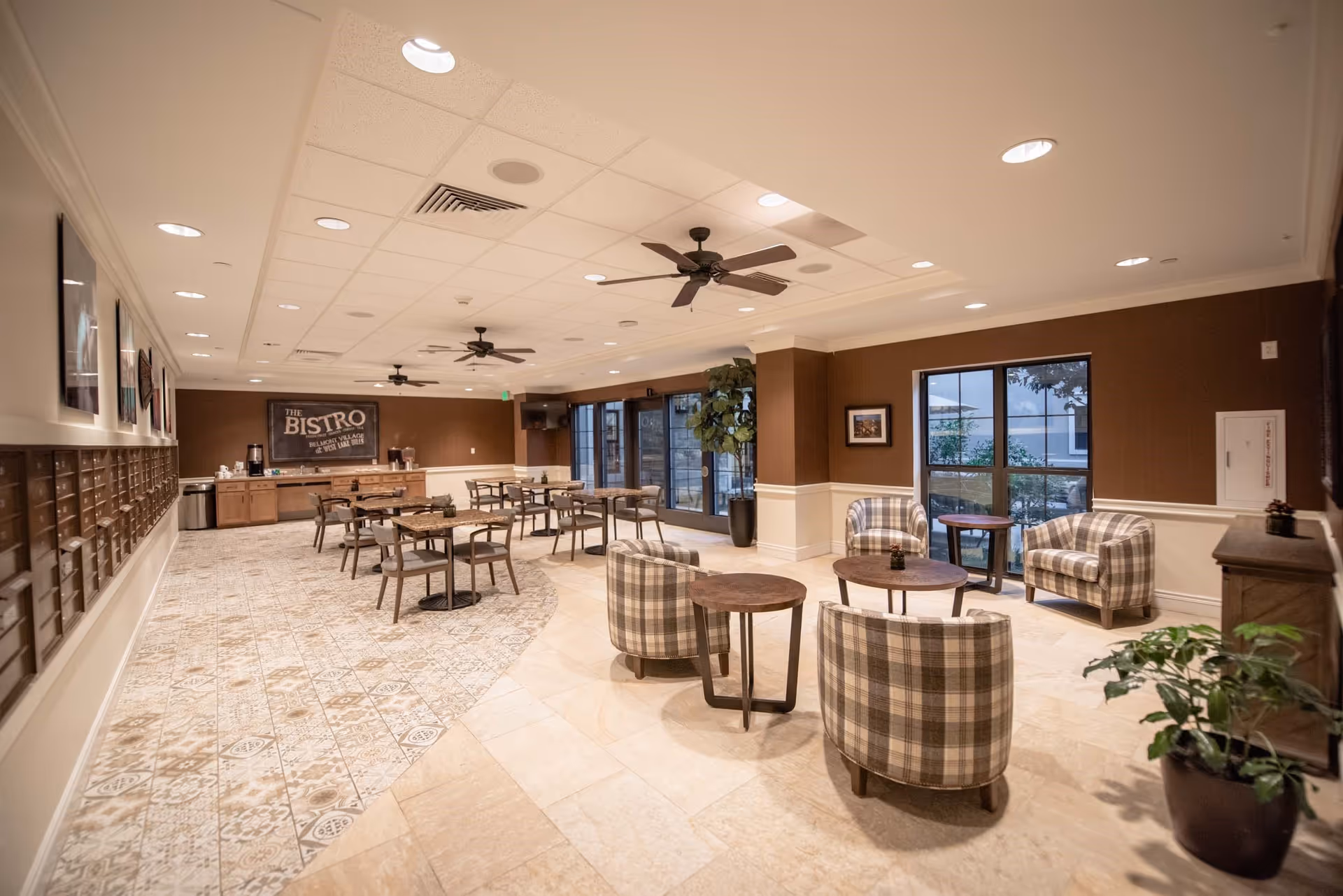 A spacious senior living facility common area with multiple tables and chairs arranged for dining and socializing. The room features patterned tile flooring, ceiling fans, large windows, and warm brown and beige walls. A sign on the back wall reads 'The Bistro'.