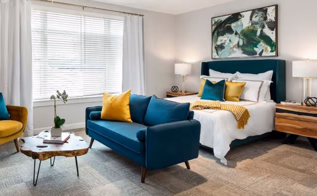 A bright and modern bedroom featuring a teal upholstered bed with white bedding and yellow and teal accent pillows. In front of the bed is a matching teal loveseat with a yellow pillow. To the left, there is a mustard yellow armchair and a rustic wooden coffee table with a potted plant and books. The room has a large window with white blinds and curtains, two wooden nightstands with lamps, and an abstract painting above the bed.