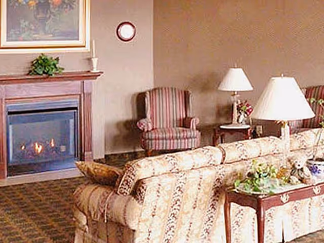 Cozy living room with a patterned sofa, a wooden side table with a lamp and decorative items, a striped armchair, and a fireplace with a wooden mantel and a painting above it.