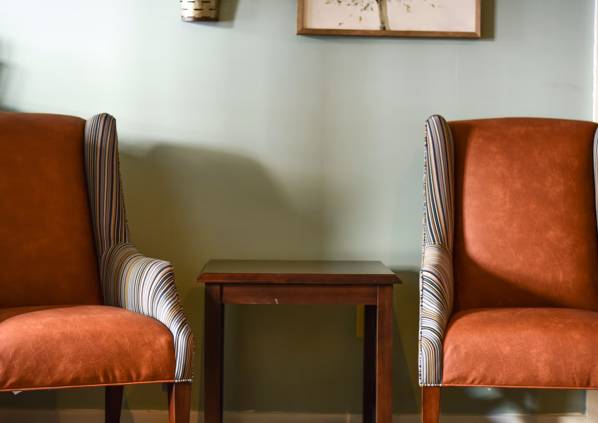 Two brown leather armchairs with striped fabric on the inner sides of the armrests, positioned on either side of a small wooden side table against a light green wall with a framed picture partially visible above.