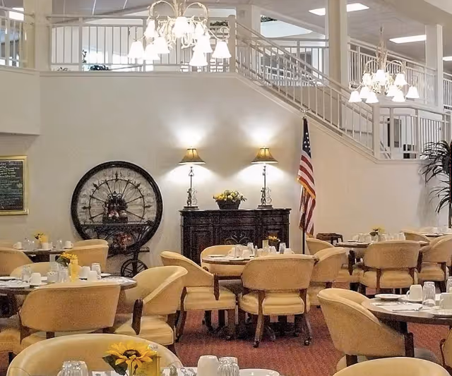 Dining room area with multiple round tables set with white dishes, glasses, and yellow flower centerpieces. Beige upholstered chairs surround the tables. In the background, there is a dark wooden sideboard with two lamps and a flower arrangement, an American flag, a large decorative wheel, and a staircase with white railings leading to an upper level.