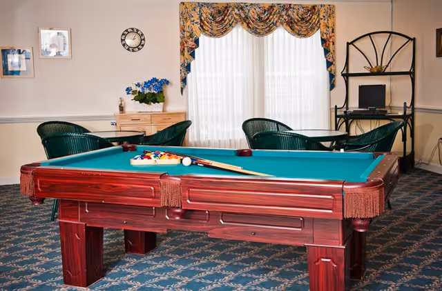 A recreational room with a wooden pool table in the center, complete with pool balls and cues. Behind the pool table, there are two round tables with green wicker chairs. A window with floral valance curtains lets in natural light. On the right side, there is a black metal shelving unit with a computer on it. The room has patterned carpet and light-colored walls with framed pictures and a clock.