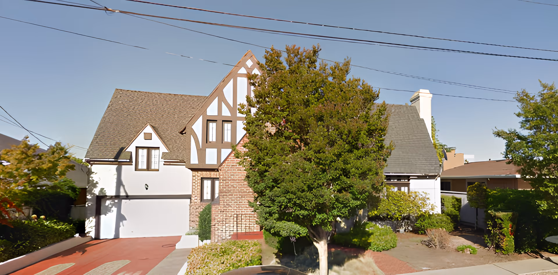 Front exterior view of a Tudor-style house with a steeply pitched roof, white walls with brown wooden trim, a brick chimney, and a large tree partially obscuring the front. There is a driveway leading to a garage on the left side and some bushes and plants around the property under a clear blue sky.