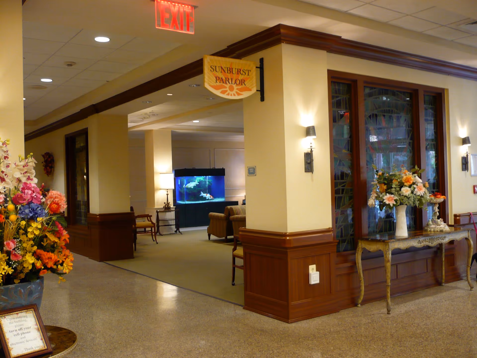 Interior view of a nursing home hallway with a sign reading 'Sunburst Parlor' hanging from the ceiling. The hallway features a large fish tank, comfortable seating, floral arrangements on a decorative table, and warm lighting fixtures on the walls.