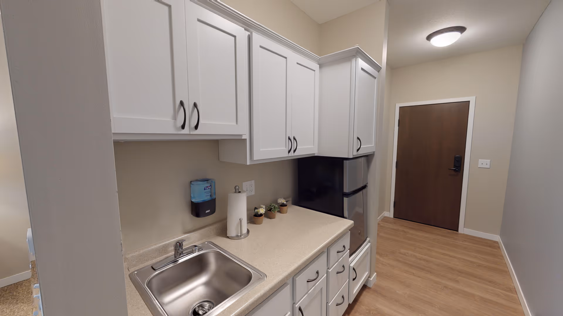 A small kitchenette area with white cabinets, a stainless steel sink, a paper towel holder, a soap dispenser, and a small refrigerator. The floor is wood, and there is a closed wooden door at the end of the hallway.