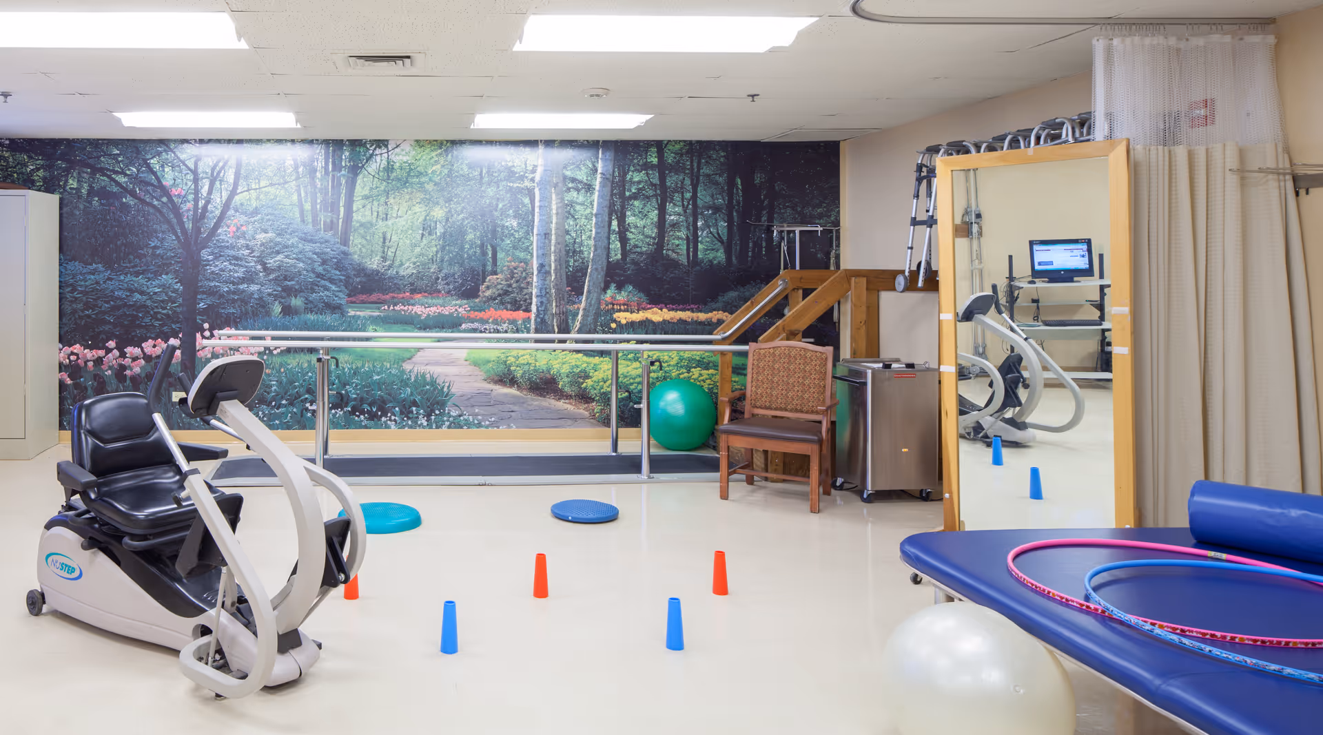 A physical therapy or exercise room with a recumbent exercise bike, colorful cones on the floor, balance pads, exercise balls, hula hoops, a large mirror, a chair, and a mural of a garden path with trees and flowers on the back wall.