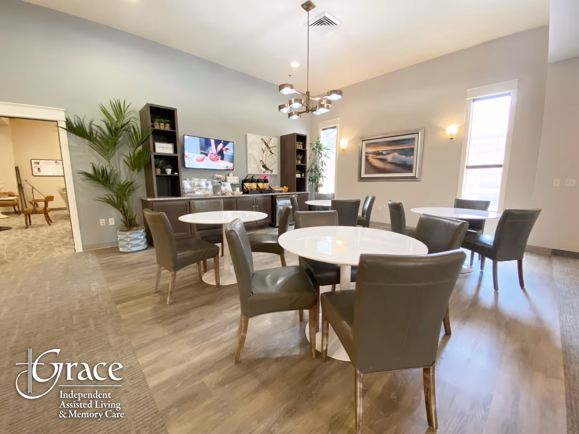 A bright and modern dining area with several round white tables surrounded by dark cushioned chairs. The room features wood flooring, a large potted plant, a wall-mounted TV, decorative artwork, and large windows letting in natural light. A chandelier hangs from the ceiling, and there is a sideboard with snacks and drinks.