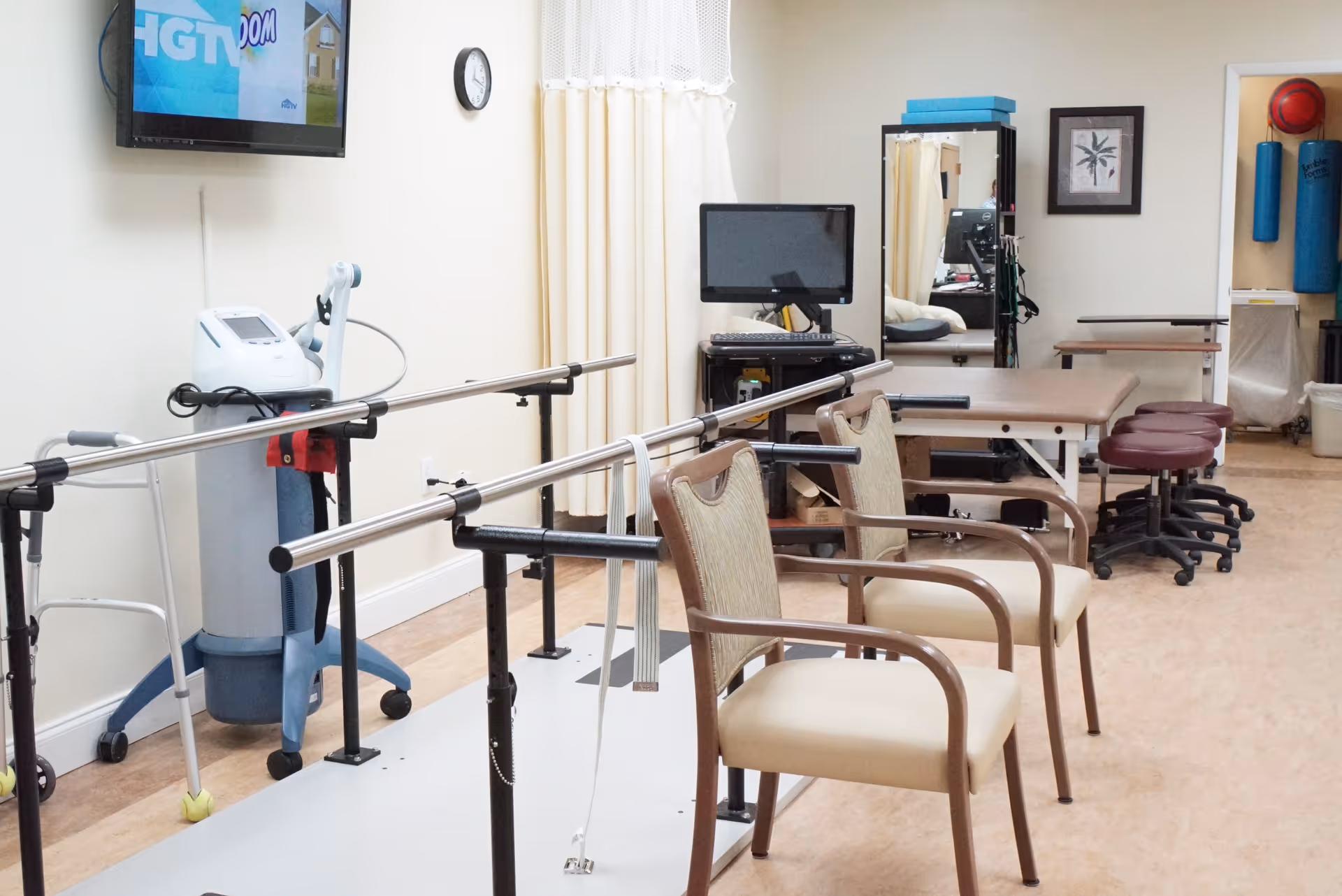 Physical therapy room with parallel bars for walking exercises, two beige chairs, a walker, a medical device, a computer on a cart, stools, and a treatment table. There is a wall-mounted TV showing the HGTV logo, a clock, and some exercise equipment in the background.
