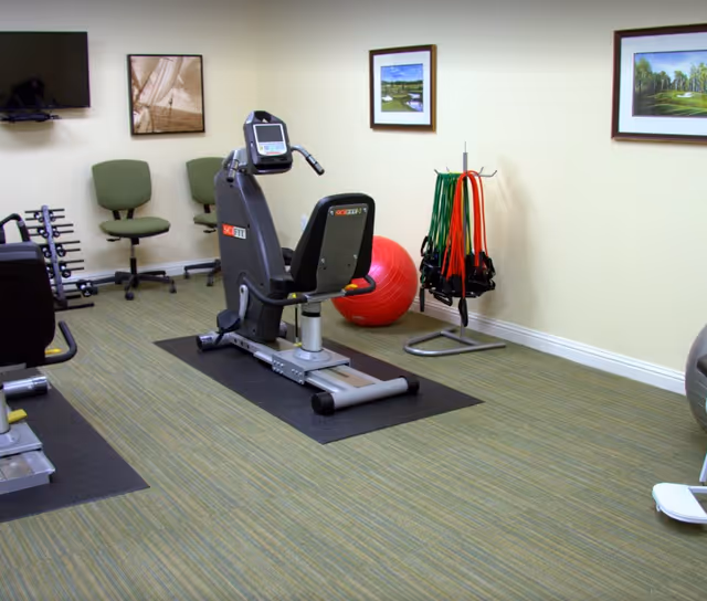 Small fitness room with a recumbent exercise bike, stability ball, resistance bands, dumbbells and chairs beneath framed wall art.