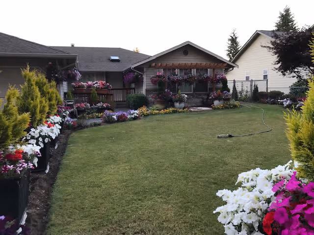 A well-maintained grassy backyard area of a residential care facility with numerous colorful flower pots and hanging flower baskets decorating the perimeter. The building has a porch with wooden railings and a small awning above the window. There are neighboring houses visible in the background.