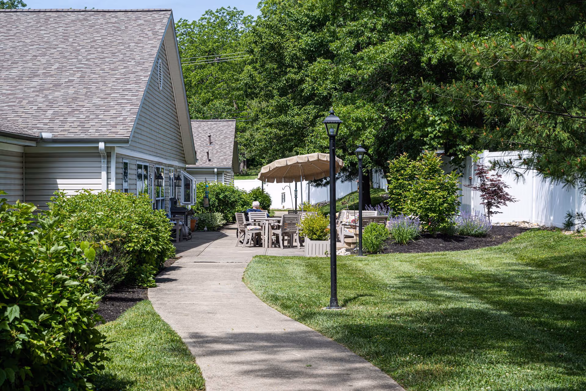 Outdoor patio area at Arden Courts - ProMedica Memory Care Community (Kenwood) with a paved walkway, green lawn, shrubs, trees, and a seating area with tables, chairs, and an umbrella. The side of a building is visible on the left.