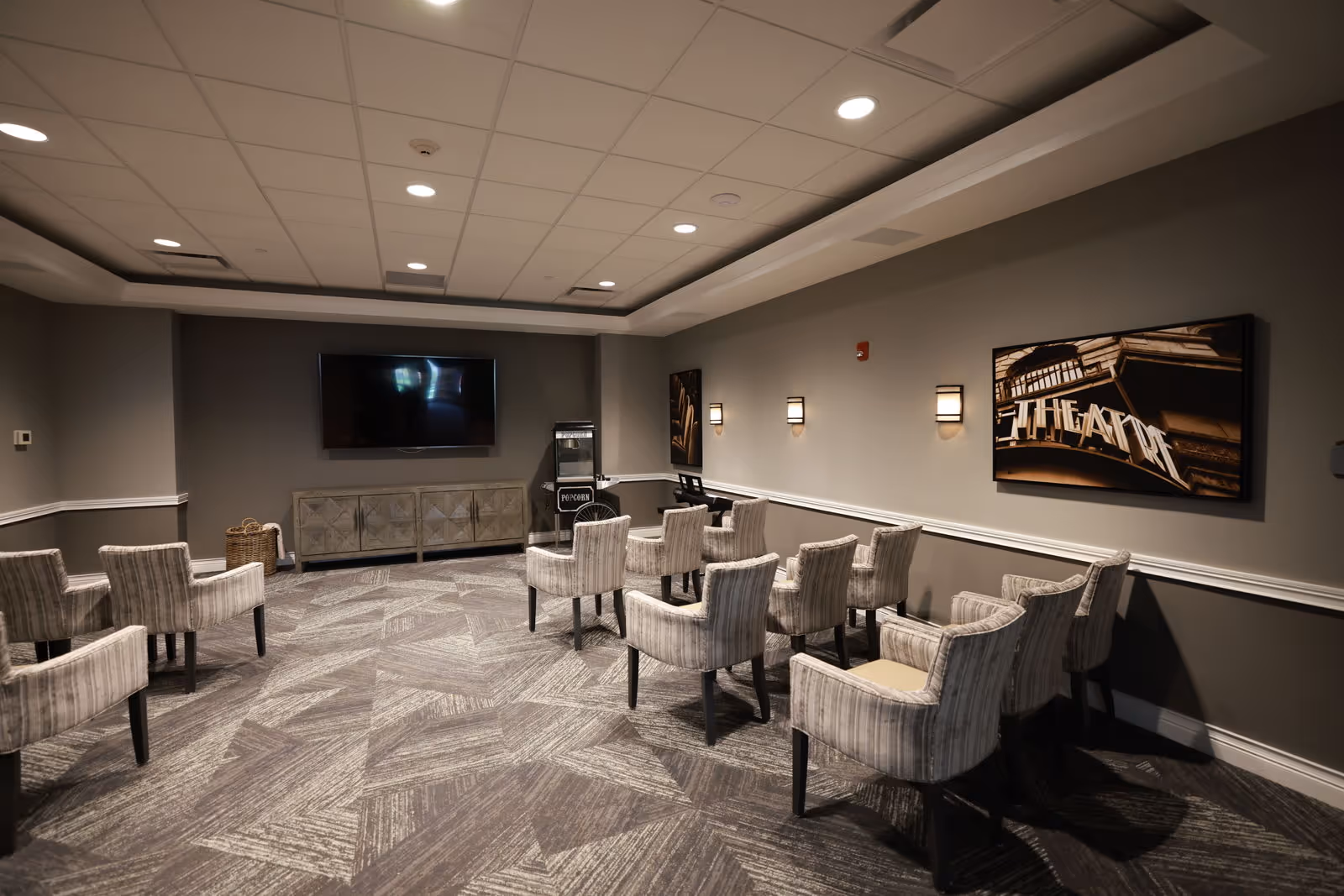 A small theater room with multiple upholstered chairs arranged in rows facing a large flat-screen TV mounted on a gray wall. There is a popcorn machine next to a cabinet beneath the TV. The room has patterned carpet flooring, soft wall lighting, and framed artwork depicting a theater marquee on the wall.