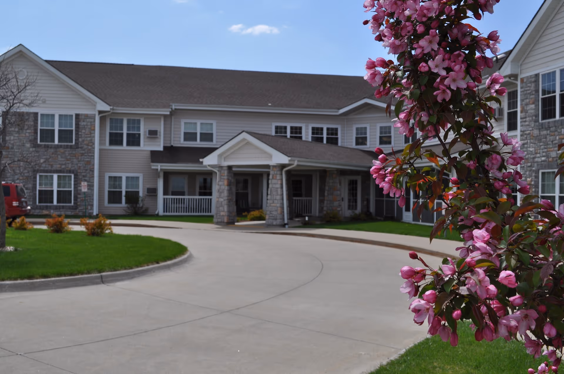 Exterior view of The Cedars Assisted Living facility showing a two-story building with stone and siding facade, a covered entrance with columns, a curved driveway, green lawn, and a flowering tree with pink blossoms in the foreground.