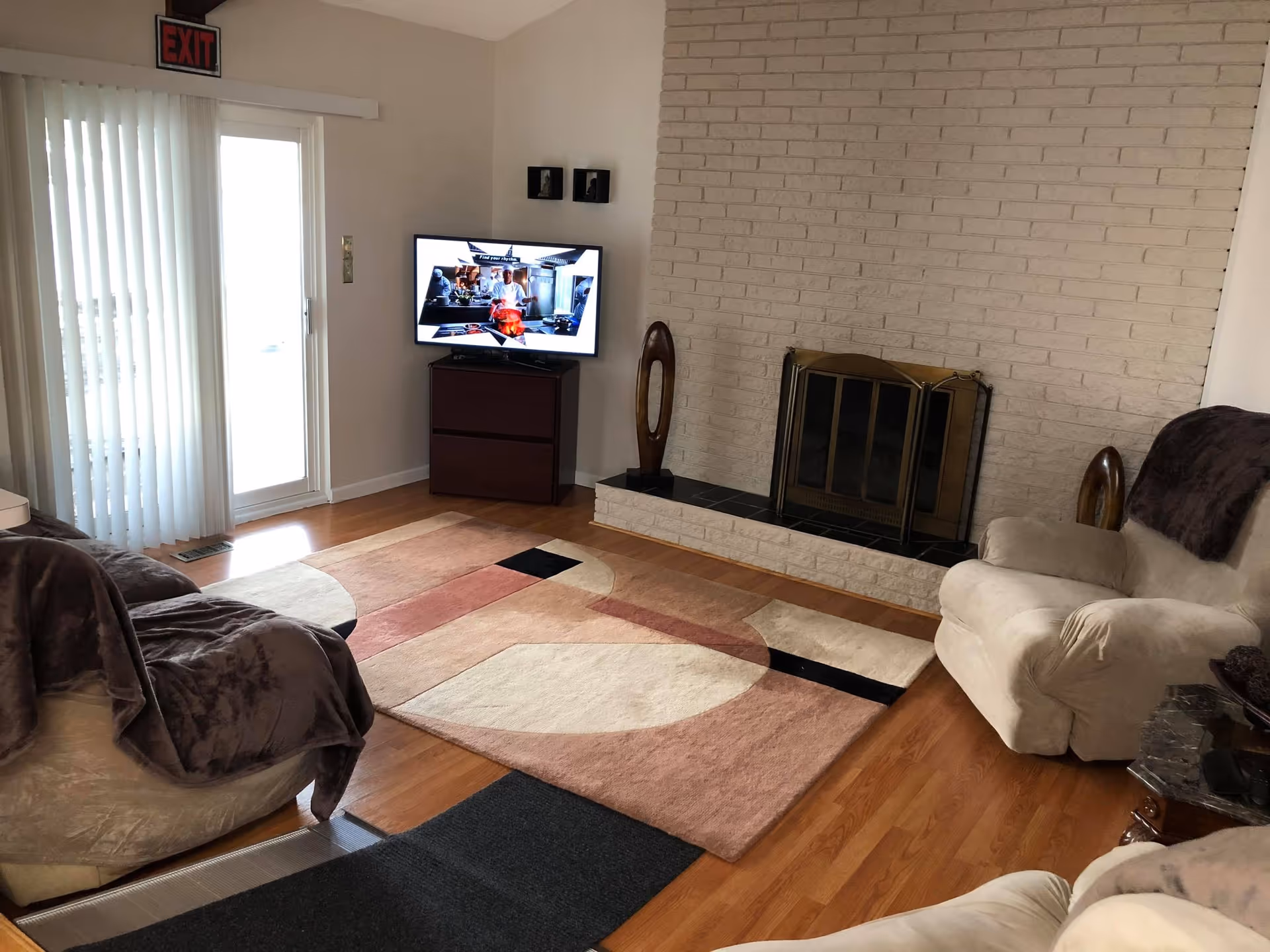 A cozy living room with wooden flooring, a beige armchair with a dark throw, a matching sofa with a dark blanket, a patterned area rug in shades of beige, pink, and black, a white brick fireplace with a brass screen, a flat-screen TV on a dark wooden stand showing a cooking show, and sliding glass doors with vertical blinds leading outside. There is an exit sign above the sliding doors.