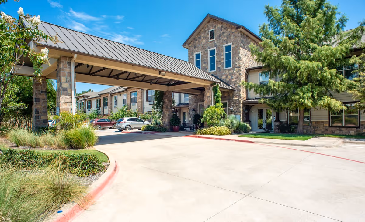 Exterior view of a senior living facility with a covered entrance supported by stone pillars, surrounded by greenery and parked cars under a clear blue sky.