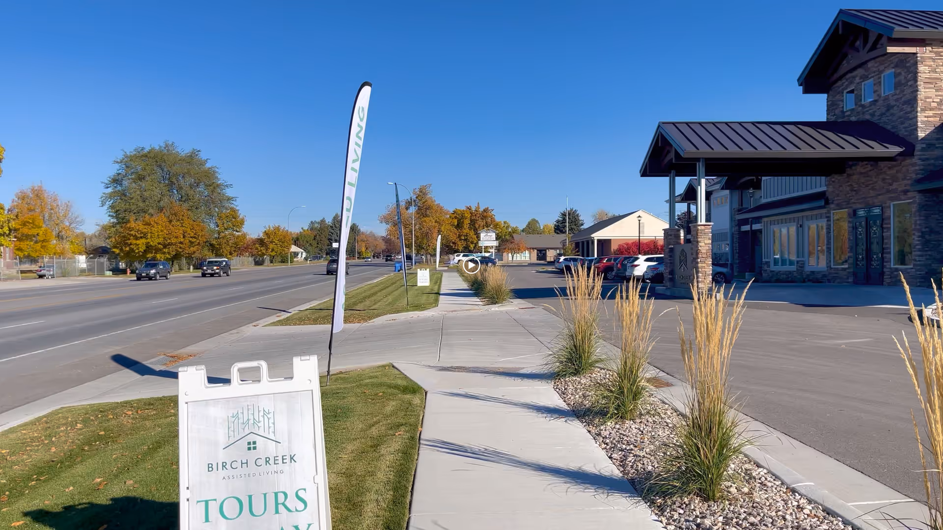 Sidewalk leading to the entrance of Birch Creek Assisted Living of Smithfield with a sign advertising tours. The building has stone and wood exterior with a covered entrance. There are ornamental grasses planted along the sidewalk and a road with cars and trees with autumn foliage in the background under a clear blue sky.