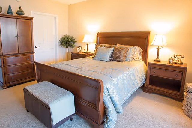 A well-lit bedroom with a wooden sleigh bed dressed in light blue bedding, matching nightstands with lamps, an armoire, and a small bench at the foot of the bed.