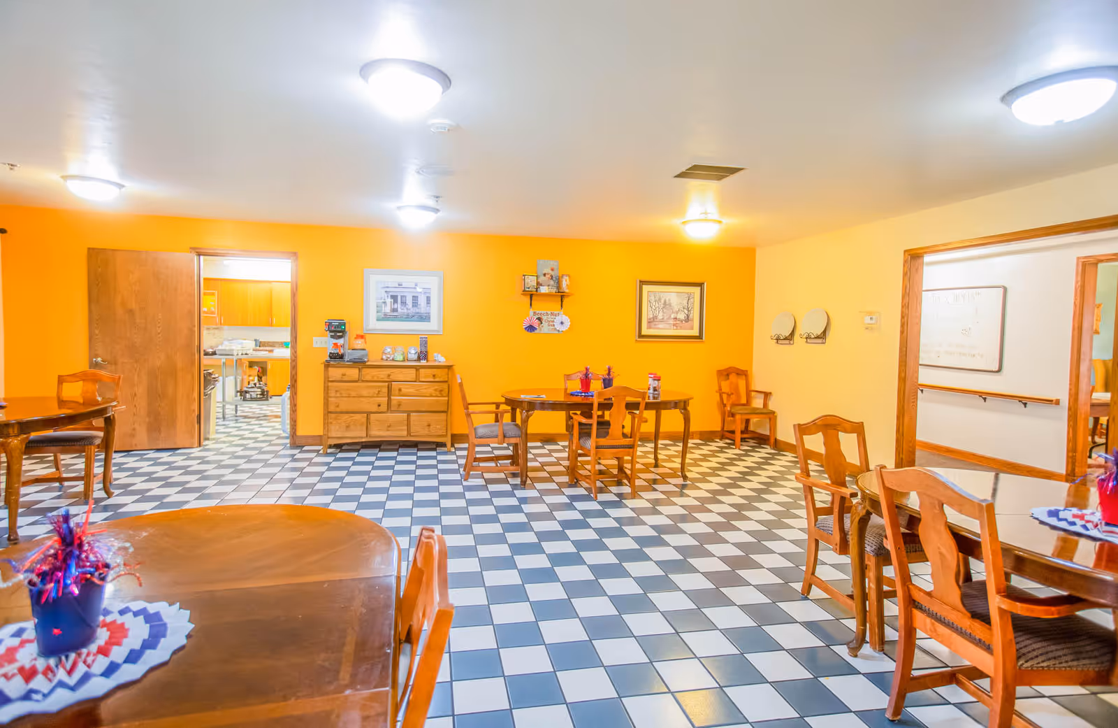 A brightly lit dining room with yellow and orange walls, checkered black and white tile floor, wooden tables and chairs, and framed pictures on the walls. There is a doorway leading to a kitchen area in the background.