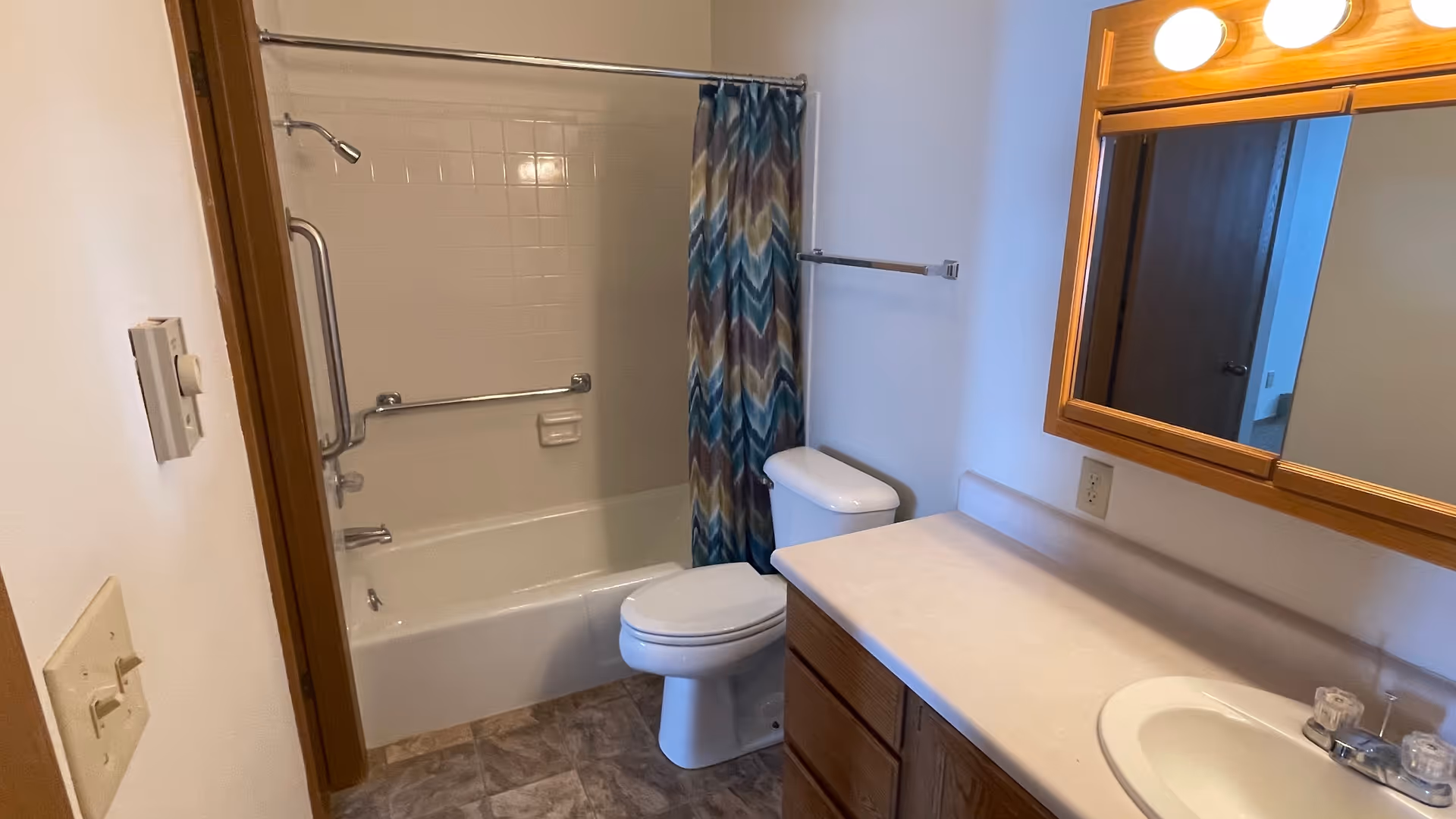 A bathroom with a white bathtub and shower combination featuring a colorful chevron-patterned shower curtain. There are two metal grab bars installed on the bathtub wall. A white toilet is positioned next to the bathtub. A wooden vanity with a white countertop and a built-in sink is on the right side, with a wooden framed mirror and two round lights above it. The floor has a tile pattern, and the walls are painted white.