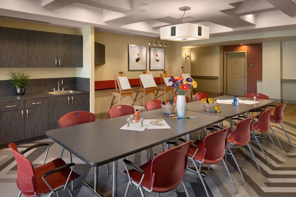 A well-lit activity room with a long table surrounded by red chairs. The table has art supplies, including markers, crayons, glue, and paper. In the background, there are easels with blank canvases and a countertop with a sink and cabinets. The room has modern lighting and framed artwork on the walls.