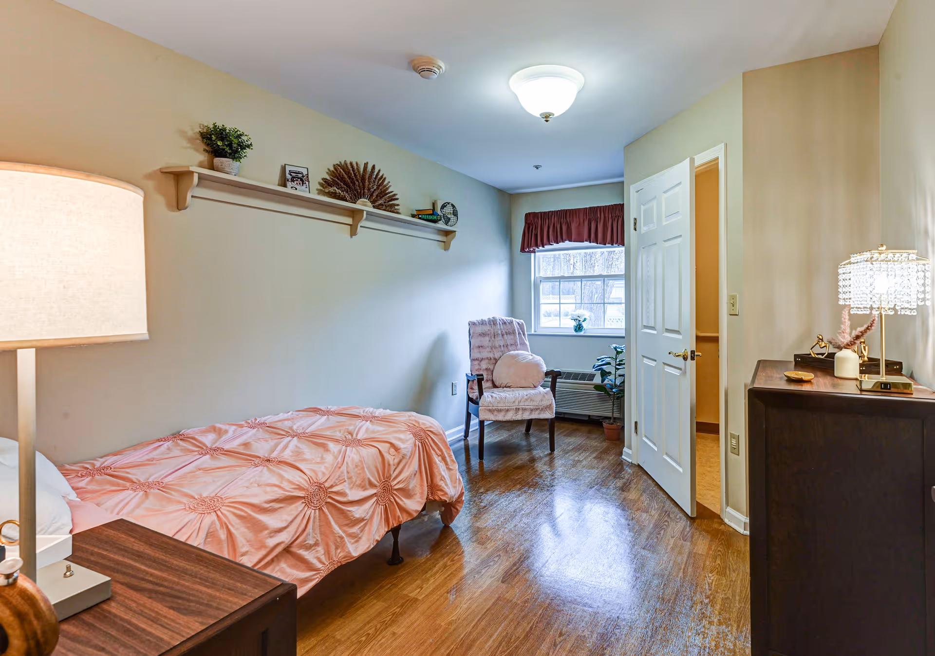 A small, cozy bedroom with a single bed covered in a pink textured bedspread. There is a wooden nightstand with a lamp on the left side of the bed. A wooden shelf mounted on the wall above the bed holds decorative items including a small plant, framed photo, and other ornaments. At the far end of the room, there is a window with a red valance, a cushioned chair with a round pillow, and a potted plant on the floor. To the right, a door is open revealing a glimpse of an adjacent room. A dark wooden dresser with a decorative lamp and small items is positioned against the right wall. The floor is wooden and the walls are painted a light beige color.