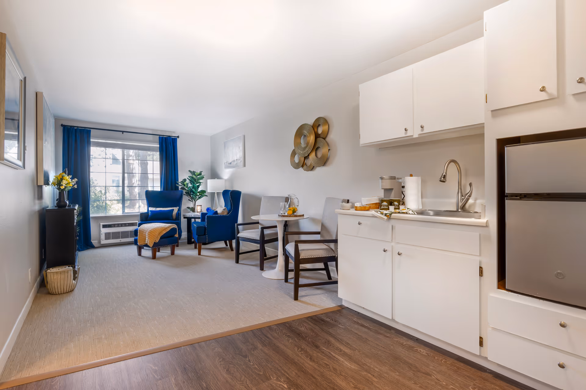 A bright and cozy living area with a small kitchenette. The kitchenette has white cabinets, a sink, and a mini refrigerator. Adjacent to the kitchenette is a small round table with two chairs. The living space features two blue upholstered armchairs with a matching ottoman, a side table with a lamp, and a large window with blue curtains letting in natural light. The walls are decorated with a metallic circular wall art and a framed picture.