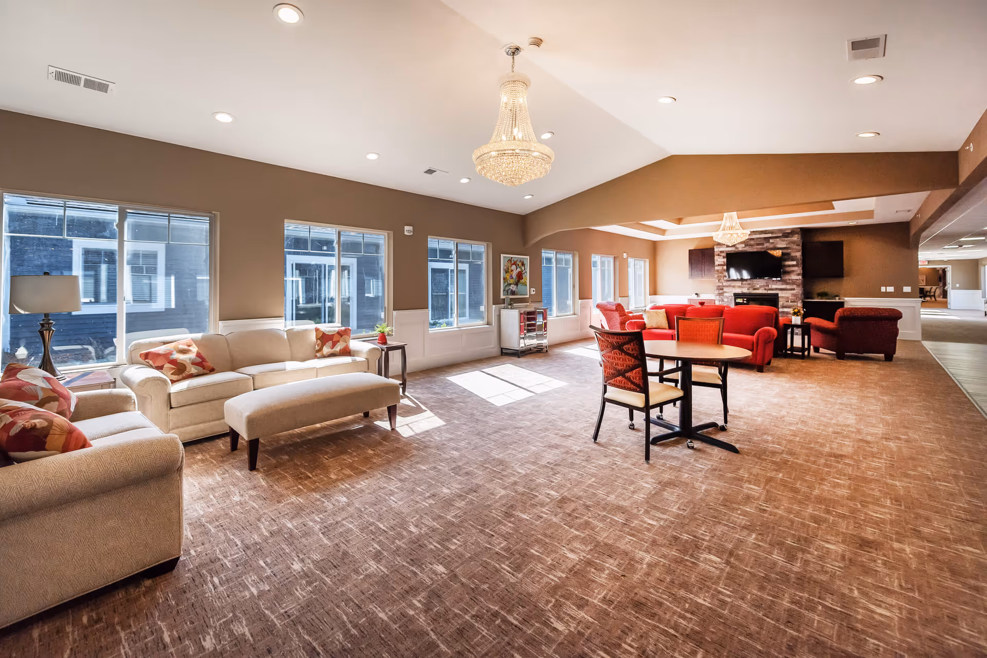 A spacious and well-lit living room area in a senior living facility featuring beige and red upholstered sofas, a matching ottoman, a round table with chairs, large windows allowing natural light, a chandelier, and a stone fireplace with a mounted flat-screen TV.