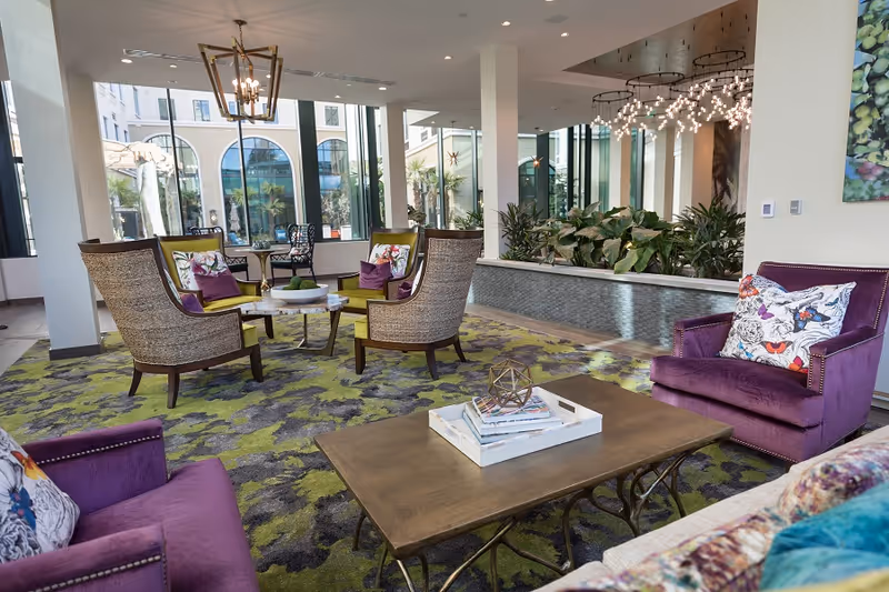 A modern and stylish living room area with purple armchairs, wicker chairs with green cushions, a wooden coffee table with a decorative tray and books, and a patterned green and gray carpet. Large windows allow natural light to fill the space, and there are plants and a unique chandelier in the background.