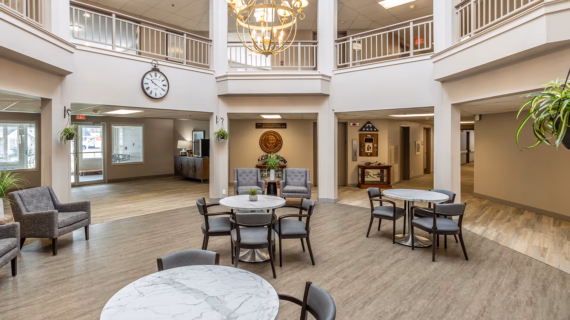 Spacious and well-lit common area in a senior living facility with round marble-top tables and chairs arranged for seating. Comfortable armchairs are placed along the walls. The area has a high ceiling with a balcony railing above and a large decorative chandelier hanging in the center. There are plants hanging from the ceiling and placed on tables, and a large clock is mounted on the wall above an open hallway.