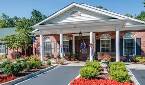 Front exterior view of a single-story brick building with white columns supporting a covered entrance. The entrance is decorated with patriotic wreaths and an American flag is displayed on the left side. There are well-maintained shrubs and flower beds with red mulch in front of the building under a clear blue sky.