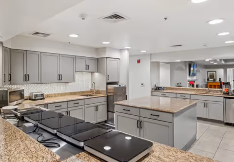 A modern kitchen with granite countertops, gray cabinets, and stainless steel appliances including a microwave and toaster. The kitchen features an island in the center and a large mirror on the wall reflecting part of the room.