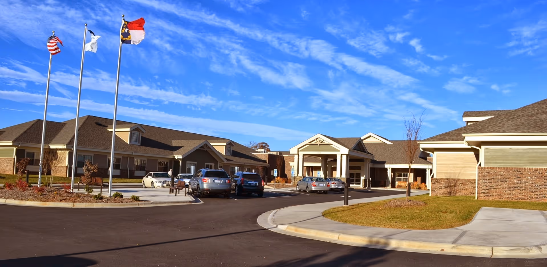Exterior view of a senior living facility named Trinity Ridge with a driveway, parked cars, three flagpoles with flags, and a clear blue sky with some clouds.