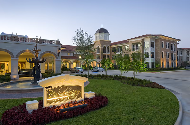 Exterior view of Conservatory At Alden Bridge senior living facility at dusk, featuring a well-lit entrance with arches, a decorative fountain, landscaped greenery, and a sign displaying the facility's name.