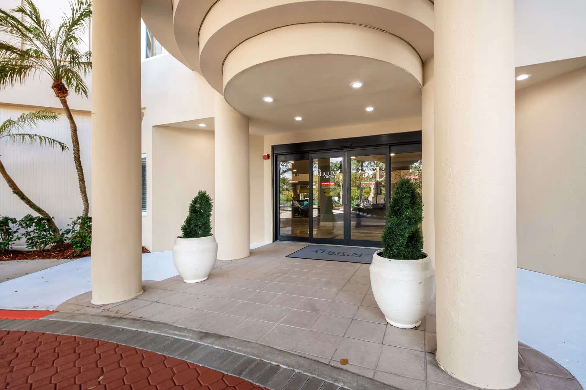 Entrance to The Atrium at Boca Raton facility showing a covered walkway with large beige columns, two white planters with green shrubs, and glass double doors with the facility name visible on them.
