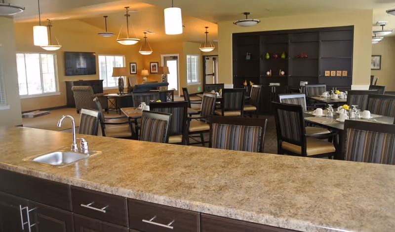 Interior view of a senior living facility dining area with multiple tables and chairs arranged neatly. The foreground shows a countertop with a small sink and faucet. The background includes a seating area with armchairs, a wall-mounted TV, and decorative shelves with items and framed pictures on the walls. The room is well-lit with ceiling lights and natural light from windows.
