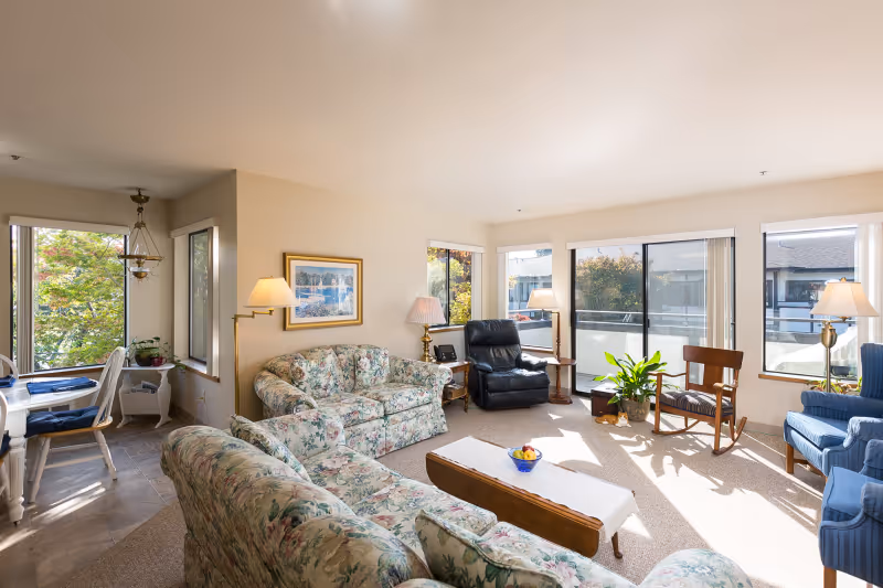 A bright and spacious living room in a senior independent living facility featuring floral patterned sofas, a black recliner, a wooden rocking chair, and blue armchairs. The room has large windows and sliding glass doors letting in natural light, with a small coffee table in the center holding a bowl of fruit. There are several lamps and a framed picture on the wall, and a dining area with a table and chairs is visible in the background.