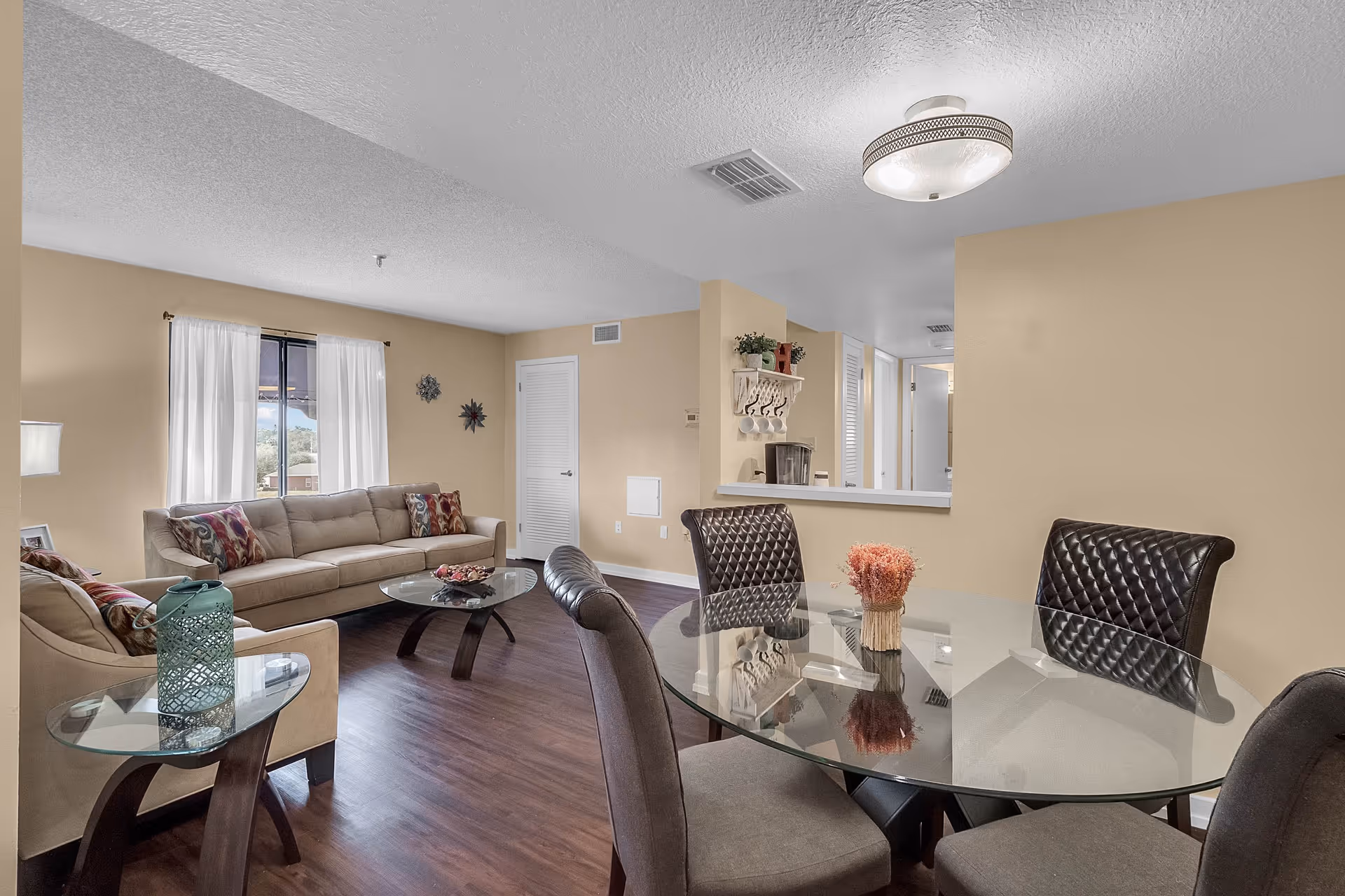 A cozy living and dining area in a senior living facility with beige walls and wood flooring. The living room features a beige sofa and armchair with colorful cushions, a glass coffee table, and a side table with a decorative lantern. The dining area has a round glass table with four cushioned chairs. A small shelf with hooks and cups is mounted on the wall near a pass-through window to the kitchen. Natural light comes through a window with white curtains.