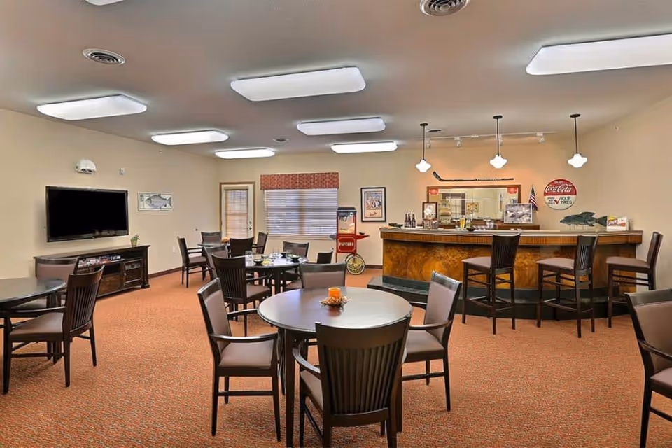 A bright senior community dining/lounge area with round tables and chairs, a wall-mounted TV, and a wooden bar counter.
