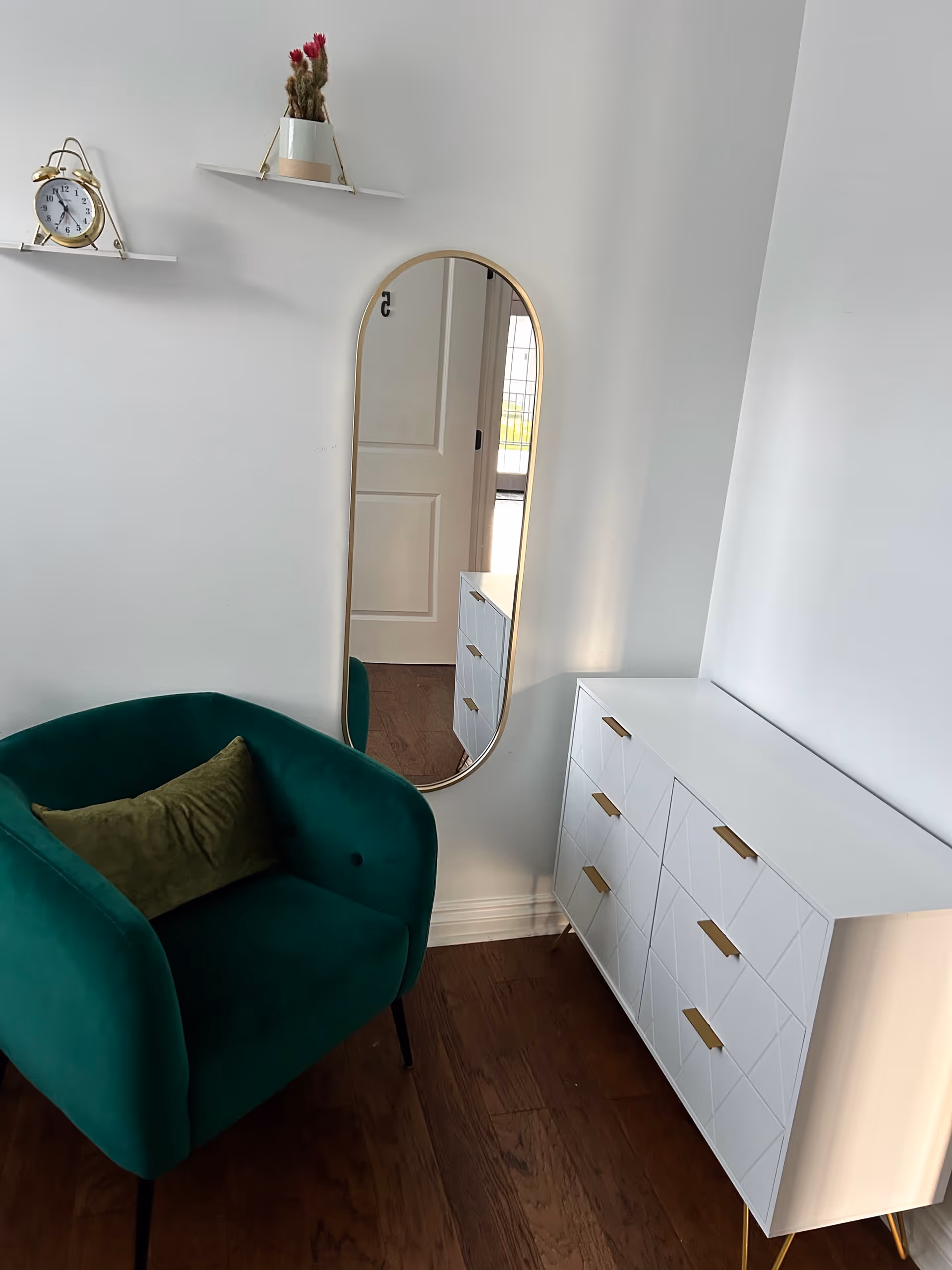 Corner of a bright room with a green velvet armchair, oval wall mirror, white dresser with gold handles, and small wall shelves.