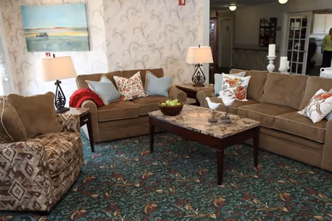Cozy living room with two beige sofas, a patterned armchair, a marble-top coffee table, lamps, and decorative pillows on a patterned carpet.