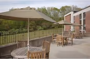 Outdoor patio area with multiple round wooden tables and chairs, each table shaded by a large beige umbrella. The patio is adjacent to a brick building with windows, and there is a metal railing along the edge overlooking a green, wooded area.