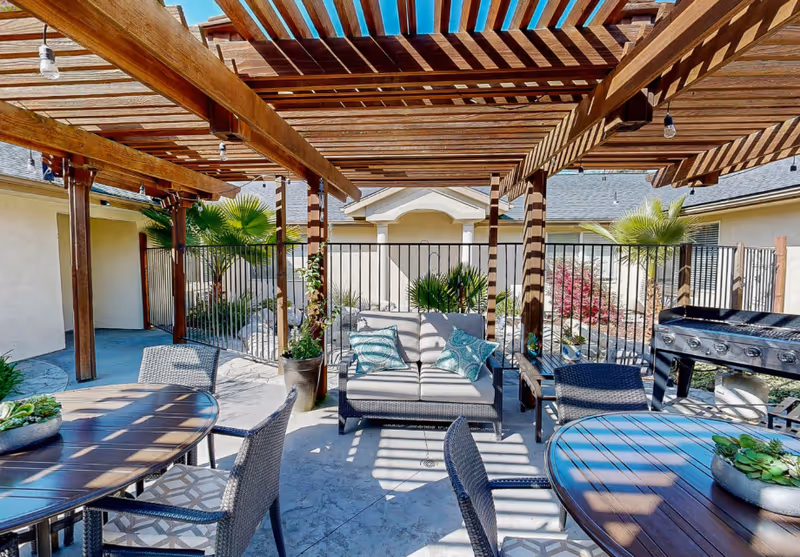 Shaded outdoor patio with a wooden pergola, wicker seating and tables, potted plants, and a barbecue grill.