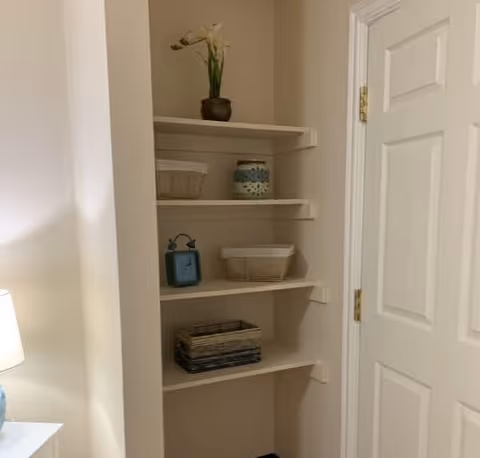 A corner of a room featuring built-in white shelves with decorative items including a potted plant, baskets, and a small clock. To the right is a white door with gold hinges, and to the left is a white table lamp on a piece of furniture.