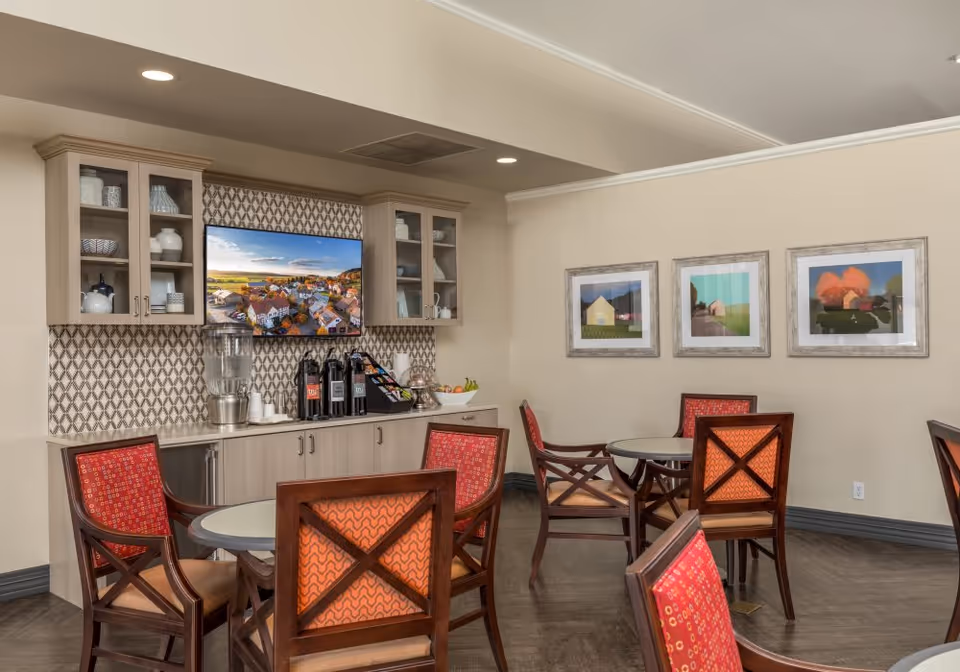 A cozy common area with round tables and wooden chairs featuring red and orange patterned cushions. On the back wall, there is a cabinet with glass doors displaying decorative items, a water dispenser, coffee urns, and a bowl of fruit. Above the cabinet, a flat-screen TV is mounted on a patterned backsplash. Three framed pictures hang on the adjacent wall.