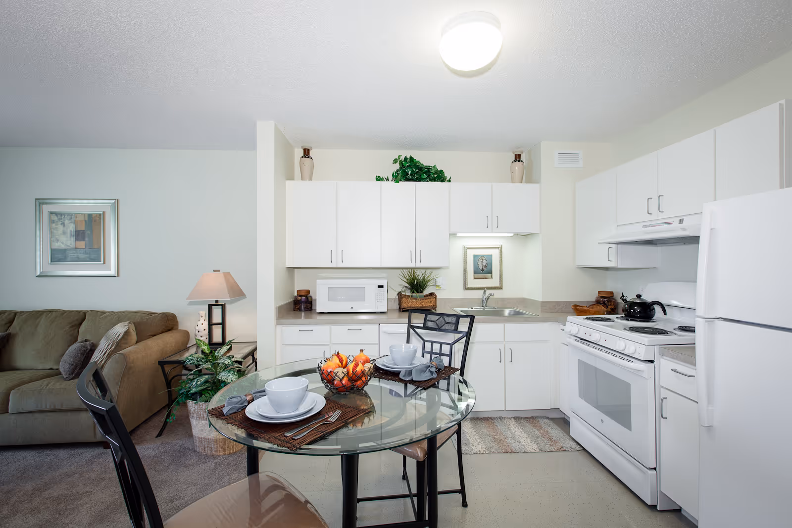 Bright open-plan kitchen and dining area adjacent to a living room, featuring white cabinets, appliances, and a glass table set for two.