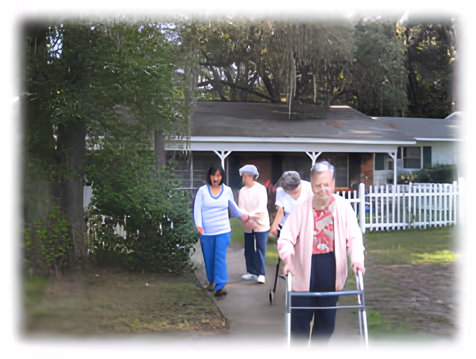 Four elderly women walking outside on a paved path near a white picket fence and a single-story house with a porch. One woman in the foreground is using a walker, while the others are walking behind her. The area is surrounded by trees and greenery.